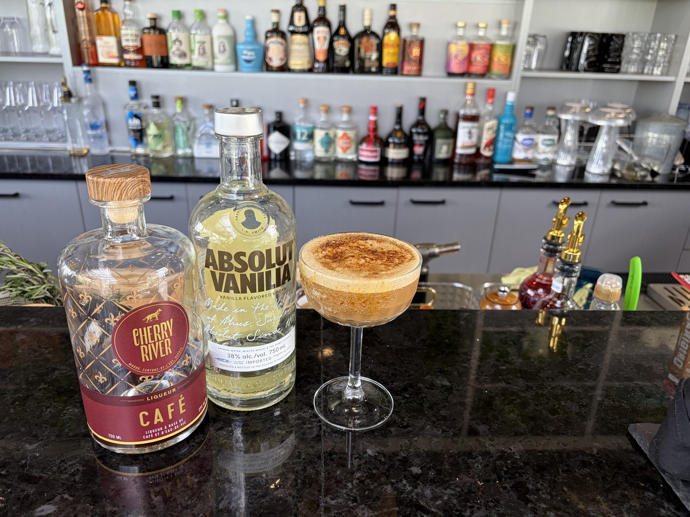 Espresso martini coiffé d'une mousse dorée, dressé au bar entre une bouteille de liqueur de café Cherry River et une vodka à la vanille